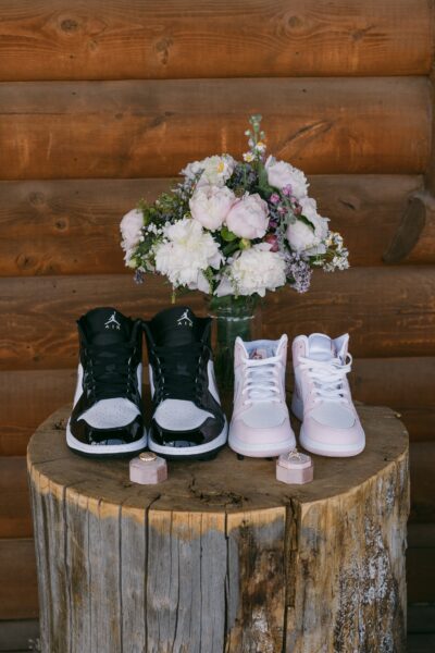 A pair of black and white Air Jordan sneakers and a pair of white and pink Air Jordan sneakers displayed on a rustic wooden stump, with a bouquet of pastel flowers in a glass vase in the background.