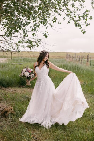 Bride in a flowing lace wedding dress, joyfully holding a bouquet of flowers and twirling in a lush green field, surrounded by trees and a rustic fence in the background.