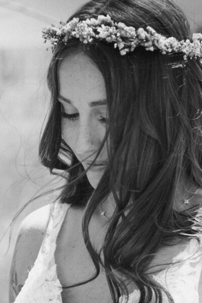 Close-up of a woman with long, flowing hair adorned with a floral crown, wearing a lace dress. She gazes downwards, surrounded by soft, natural light, capturing a serene and intimate moment.