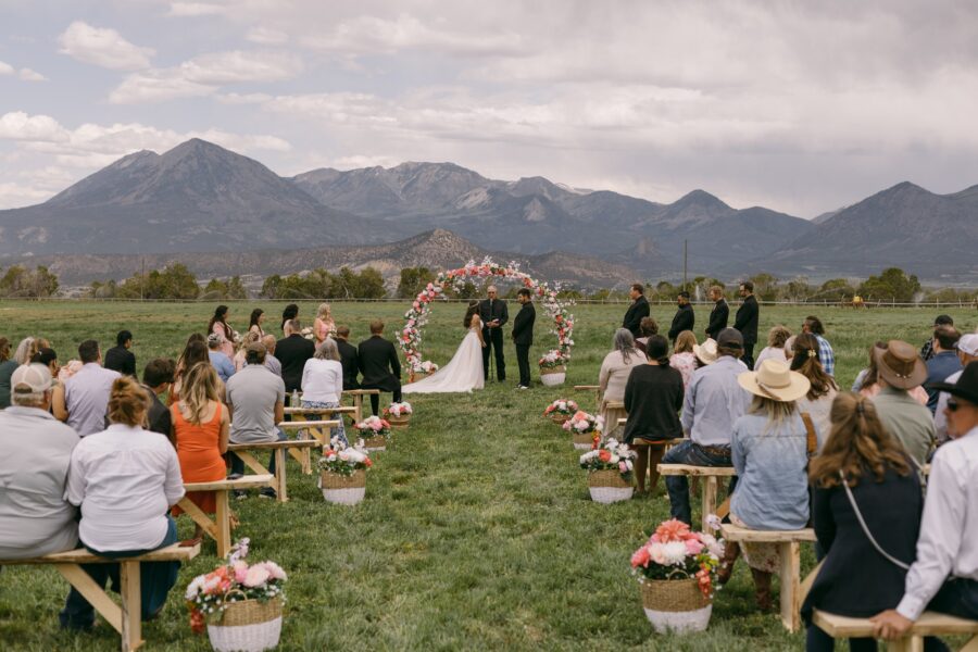 Outdoor wedding ceremony with guests seated on wooden benches, set against a backdrop of mountains. The couple stands under a floral arch while officiants and attendants look on. Bright flowers in baskets line the aisle, creating a festive atmosphere.