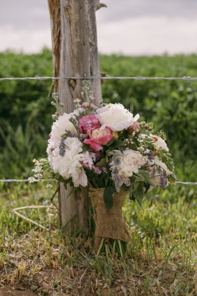 A beautiful bouquet of peonies and mixed flowers wrapped in burlap, leaning against a wooden post in a lush green field, perfect for wedding or event decor.