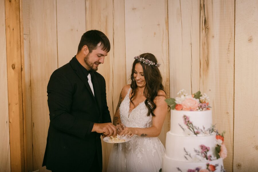 Bride and groom smiling while cutting their wedding cake, surrounded by rustic wooden walls and a beautifully decorated cake adorned with flowers.