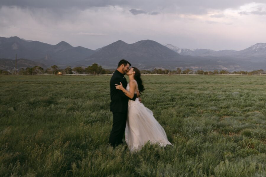 Couple embracing and sharing a kiss in a lush green field with mountains in the background, capturing a romantic wedding moment.