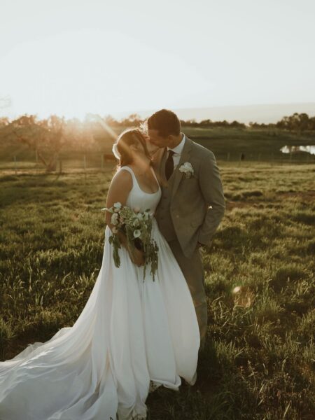 A bride and groom share a romantic kiss in a sunlit field during their outdoor wedding, with the bride wearing a flowing white gown and holding a bouquet of flowers, while the groom is dressed in a light gray suit.
