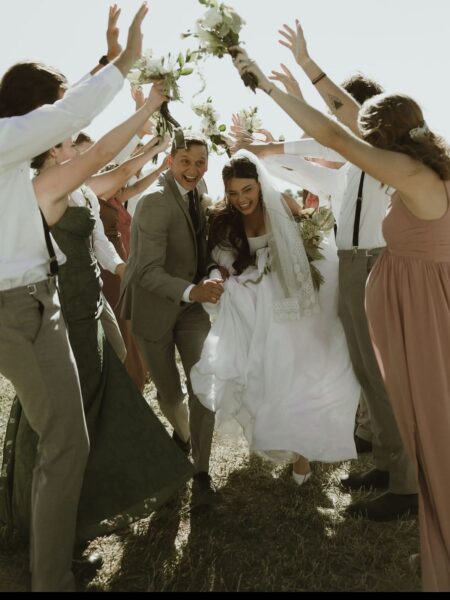 Joyful wedding celebration with the bride and groom surrounded by their wedding party, holding bouquets and cheering. The couple is laughing and running together under a floral arch, set in a sunny outdoor location.