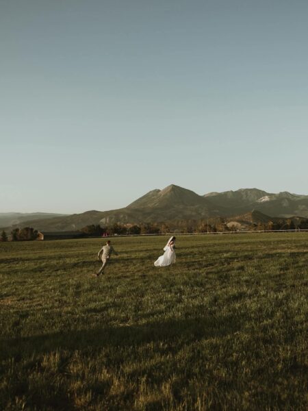 A couple in formal attire joyfully runs through a lush green field with mountains in the background, capturing a romantic and adventurous moment during their wedding celebration.