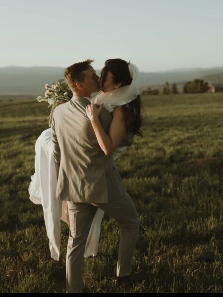 Couple sharing a romantic kiss in a scenic outdoor setting, with the groom in a light gray suit and the bride in a flowing white dress holding a bouquet, surrounded by lush green grass and mountains in the background.