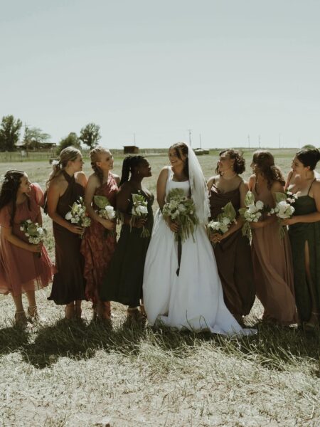 Bridal party smiling together in a field, featuring the bride in a white gown and her bridesmaids in various colorful dresses, each holding bouquets of flowers.