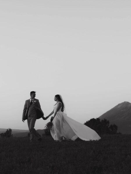 Black and white image of a bride and groom walking hand in hand on a grassy hill, with a mountain backdrop during sunset, capturing a romantic wedding moment.