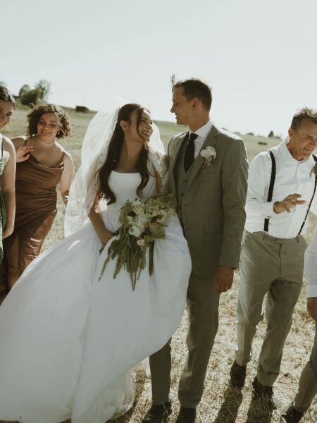 Bride and groom smiling together in a sunny outdoor setting, surrounded by wedding party members. The bride wears a white gown and veil, holding a bouquet, while the groom is dressed in a gray suit. Friends in elegant dresses and suits add to the joyful atmosphere of the celebration.