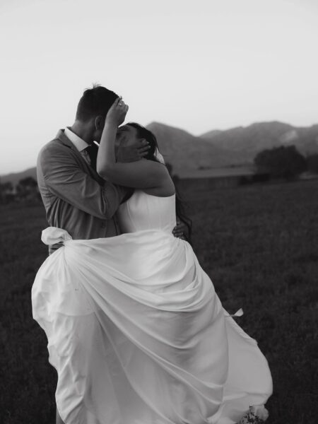 Couple embracing in a romantic kiss during their wedding, wearing elegant attire with a flowing white wedding dress, set against a picturesque mountain backdrop at sunset.