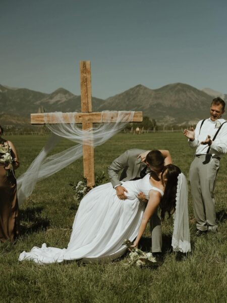 Bride and groom sharing a romantic kiss in front of a wooden cross during an outdoor wedding ceremony, surrounded by mountains and lush greenery, with bridal party members celebrating nearby.