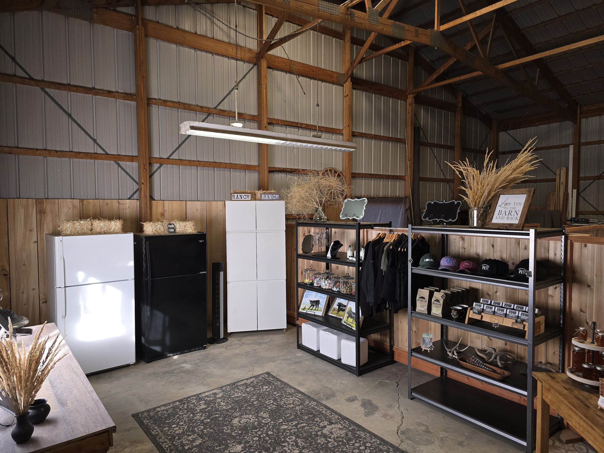 Market Interior view of a rustic barn showcasing three refrigerators, shelving units filled with merchandise, and decorative elements like dried grass and signs, highlighting a blend of functionality and country charm.