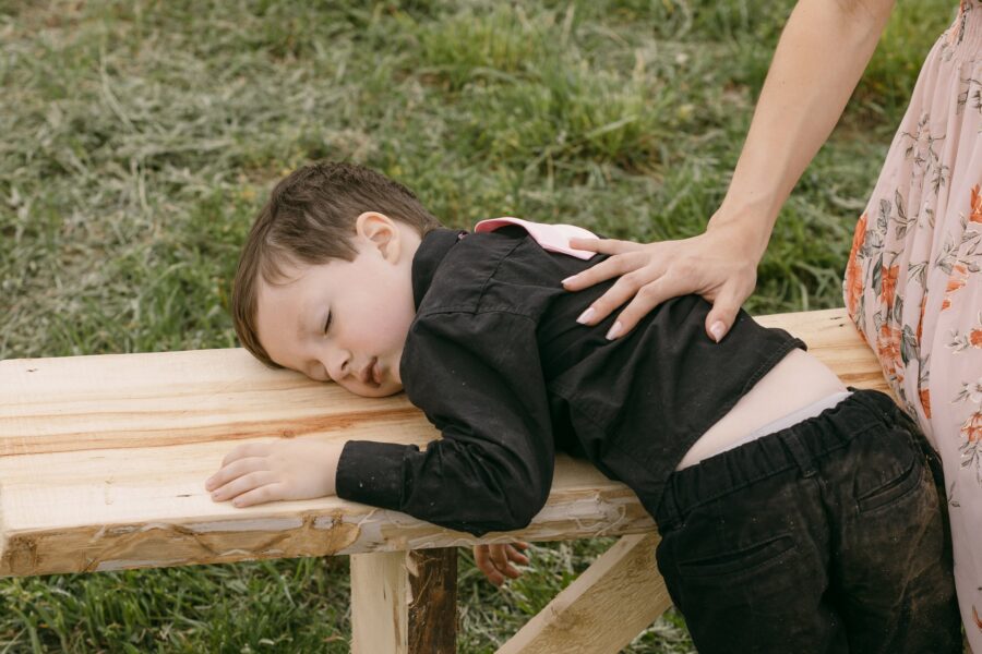 A young boy resting his head on a wooden bench, looking tired, while a woman gently places her hand on his back. The scene is set outdoors with green grass in the background, capturing a moment of care and comfort.