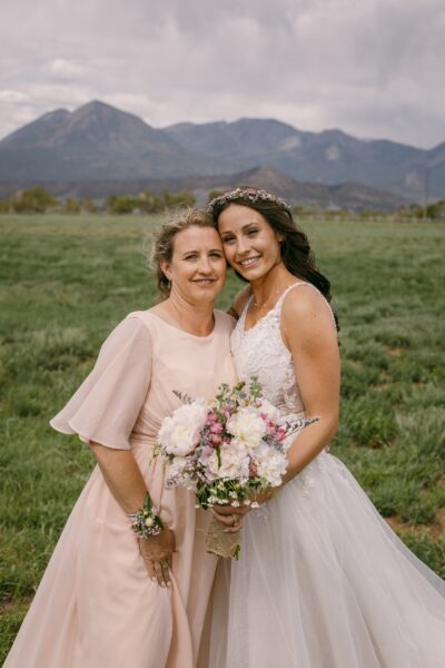 Bride and her mother pose together in a scenic outdoor setting with mountains in the background, showcasing the bride in a beautiful lace wedding gown and floral crown, while the mother wears a soft pink dress and holds a bouquet of flowers.