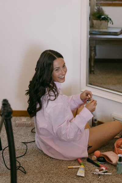 A woman with long, wavy hair sits on the floor in a cozy room, wearing a pink button-up shirt. She smiles while holding a beauty product, surrounded by various makeup items and tools scattered around her. A mirror reflects her cheerful expression, adding a personal touch to the scene.