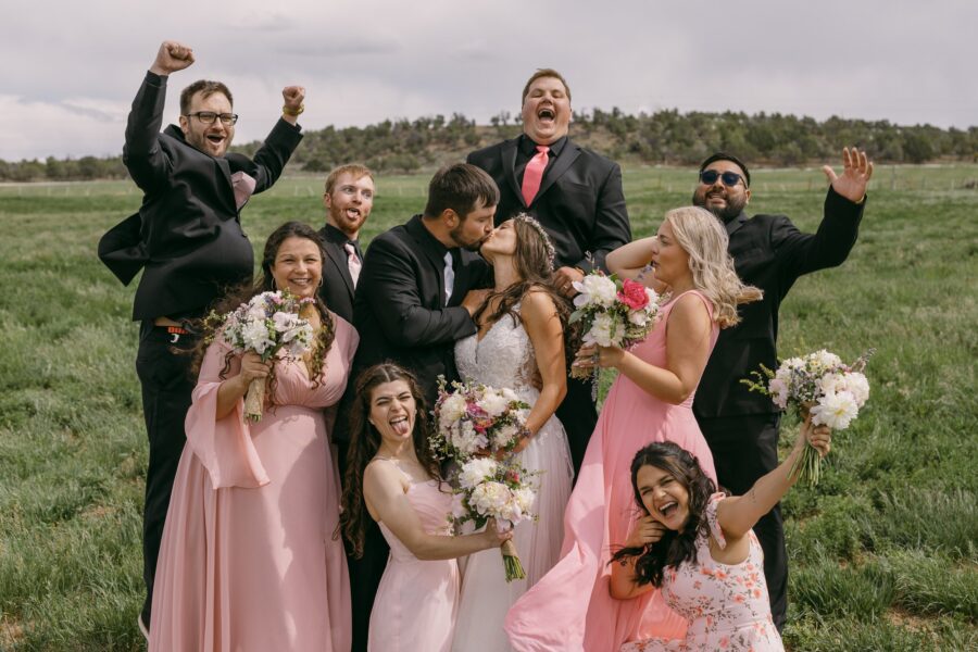 Wedding party celebrating outdoors, featuring the bride and groom in a joyful kiss surrounded by friends in formal attire and pink dresses, with bouquets of flowers, set against a lush green field.