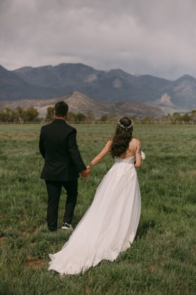 Couple holding hands while walking through a green field with mountains in the background, showcasing a romantic outdoor wedding setting. The bride wears a white wedding gown and floral crown, while the groom is dressed in a black suit.