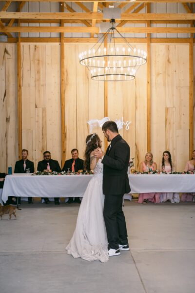 Couple sharing their first dance at a wedding reception in a rustic venue, surrounded by guests seated at a long table adorned with flowers. The setting features wooden walls, a large chandelier, and cheerful decorations, creating a romantic atmosphere.