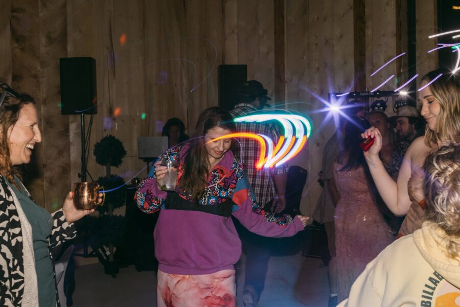 Group of friends dancing at a lively celebration, with colorful light trails and drinks in hand. The scene features a warm, rustic venue with wooden walls, showcasing joyful expressions and dynamic movement.