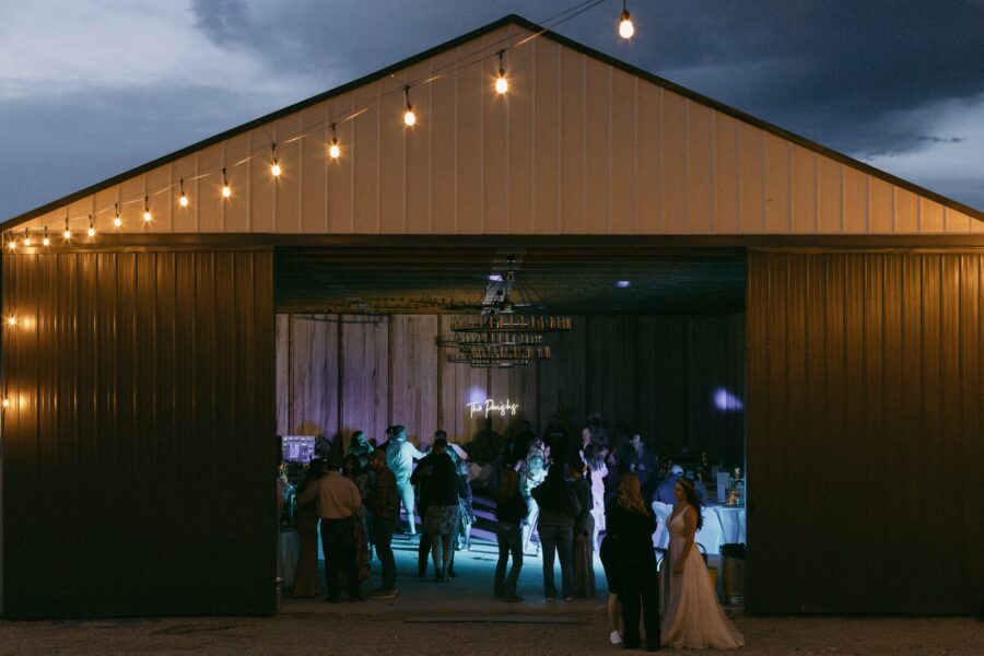 A lively indoor event taking place in a barn with string lights illuminating the space, featuring guests dancing and socializing, while a bride in a white gown stands in the foreground.