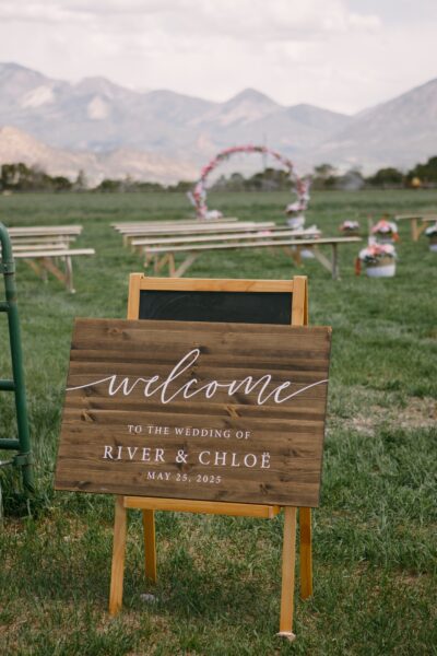 Wooden welcome sign for the wedding of River and Chloë, displayed outdoors with a scenic mountain backdrop, featuring a floral arch and seating arrangements for guests. Date: May 25, 2025.