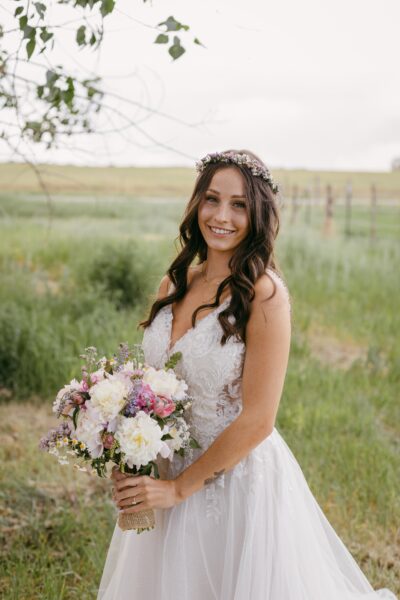 Bride in a beautiful lace wedding gown holding a vibrant bouquet of flowers, standing in a lush green field with a serene landscape in the background. Her long, dark hair is adorned with a floral crown, and she is smiling warmly at the camera.