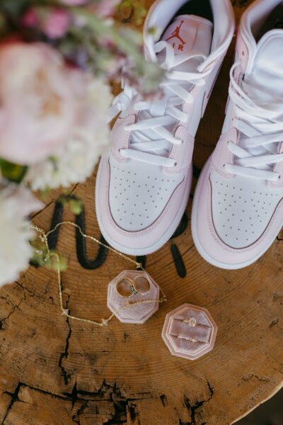 Stylish white and pink Air Jordan sneakers placed on a rustic wooden surface, accompanied by elegant gold jewelry including a ring and necklace in velvet display boxes, with fresh flowers in the background.