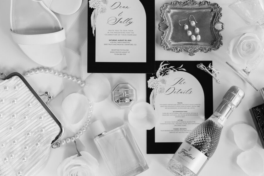 Elegant wedding invitation suite featuring details for Dean and Sally's celebration, alongside accessories like pearl earrings, a stylish clutch, a bottle of Prosecco, and a floral arrangement on a soft fabric background.