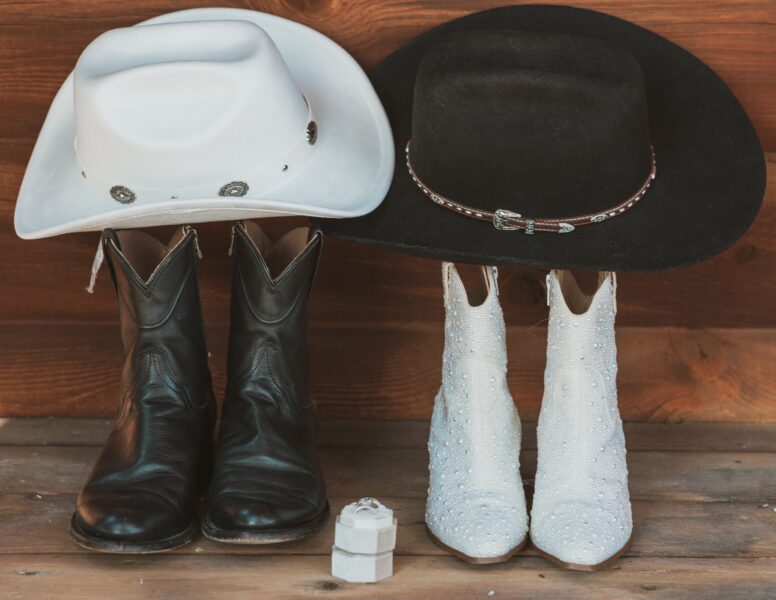Pair of cowboy boots and hats displayed on wooden surface, featuring a black felt hat with decorative band, a white felt hat, glossy black leather boots, and sparkling white ankle boots embellished with rhinestones.