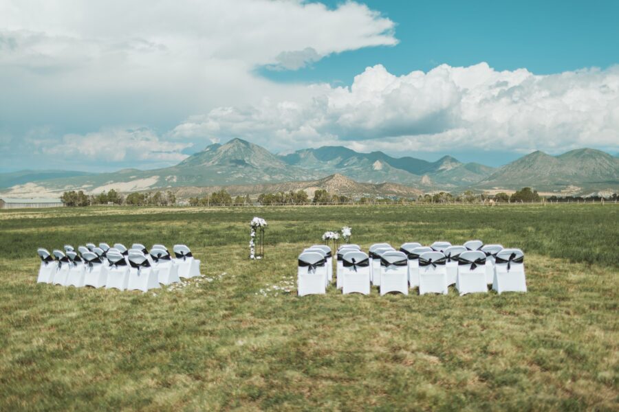 Outdoor wedding setup featuring white chairs with black sashes arranged in a circular pattern, surrounded by lush green grass and scenic mountains under a partly cloudy sky.