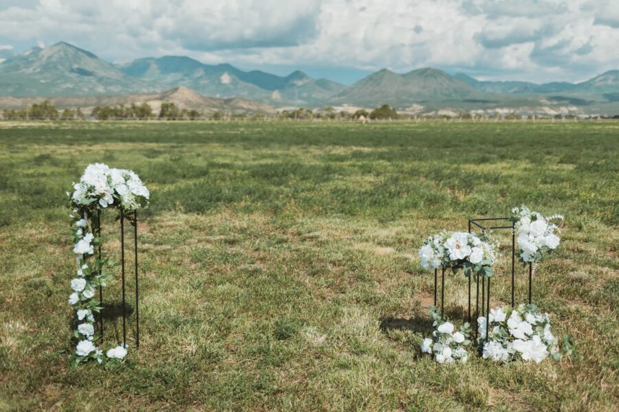 Outdoor wedding decor featuring white floral arrangements on black metal stands, set against a backdrop of mountains and a green field. Ideal for a romantic and scenic ceremony setting.