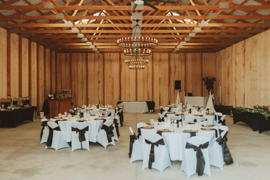 Elegant event space featuring round tables with white linens and black bows, set up for a formal gathering. The rustic wooden walls and exposed beam ceiling complement the chandeliers, creating a warm and inviting atmosphere. A bar area is visible on the left, enhancing the venue's charm.