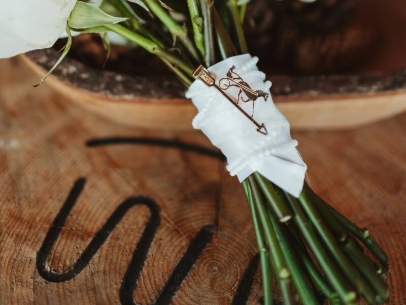 Close-up of a bridal bouquet wrapped in white satin ribbon, featuring a decorative gold pin, placed on a rustic wooden surface with visible grain patterns.