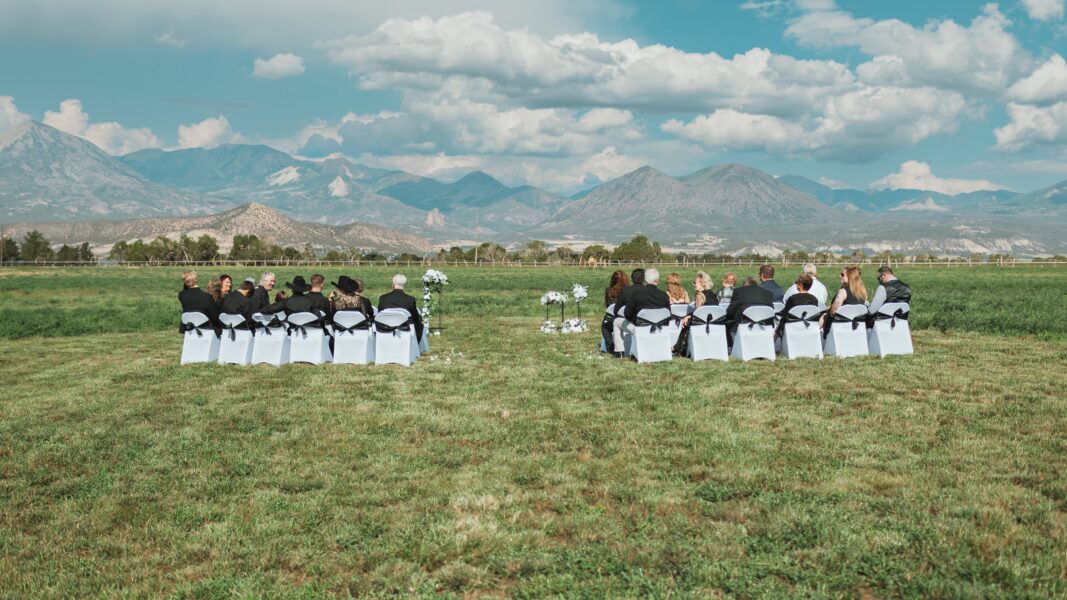 Outdoor wedding ceremony with guests seated on white chairs adorned with black ribbons, set against a picturesque mountain backdrop under a blue sky with clouds.