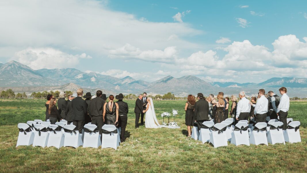 A scenic outdoor wedding ceremony with a couple exchanging vows in front of a mountain backdrop, surrounded by guests seated in white chairs adorned with black sashes. The sky is partly cloudy, and the lush green field adds to the romantic atmosphere.