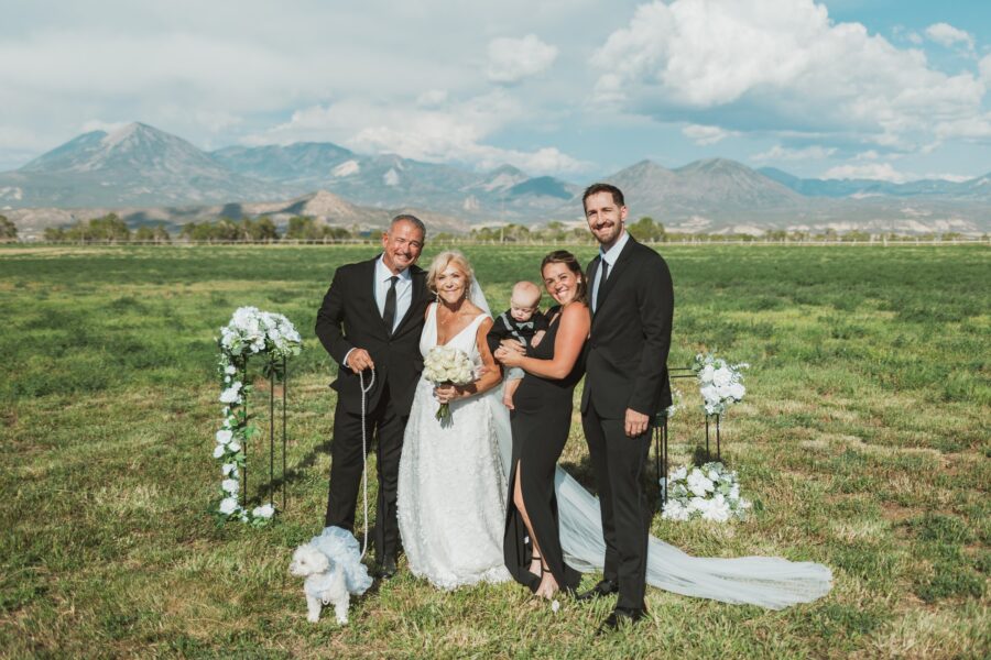 Wedding party posing outdoors with mountains in the background, featuring a bride in a white dress holding a bouquet, a groom in a black suit, a bridesmaid with a baby, and a dog in a decorative outfit. Floral arch decorations enhance the picturesque setting.