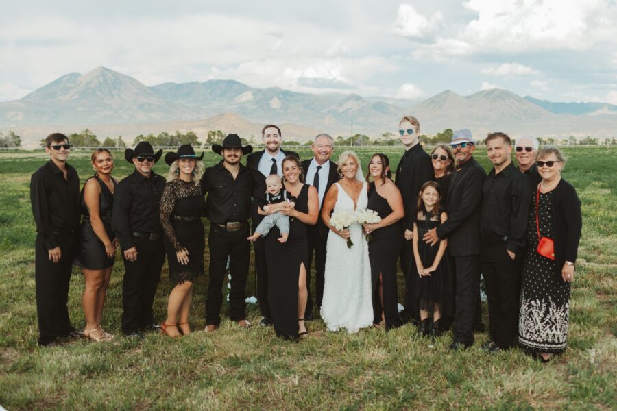Family gathering in black attire at a scenic outdoor location, featuring a bride in a white dress holding flowers, surrounded by family members of various ages, with mountains in the background.