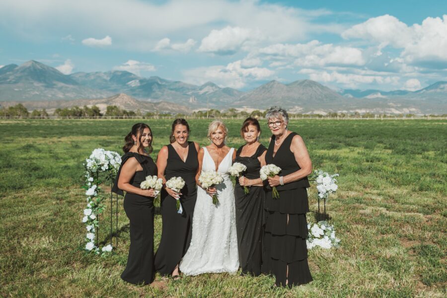 Bridal party posing outdoors in a scenic field with mountains in the background, featuring the bride in a white dress surrounded by four women in black dresses, each holding bouquets of white roses.
