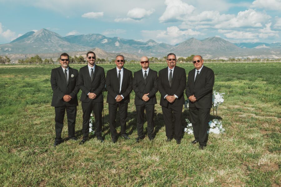 Group of six men in black suits and sunglasses standing in a grassy field with mountains in the background, ideal for a wedding or formal event setting.