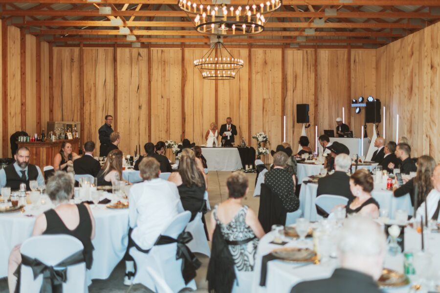 Wedding reception scene in a rustic venue with wooden walls, featuring guests seated at elegantly set tables. A couple stands at the front, with a speaker delivering a toast, while a DJ is set up in the corner. The ambiance is festive, highlighted by chandeliers and floral decorations.