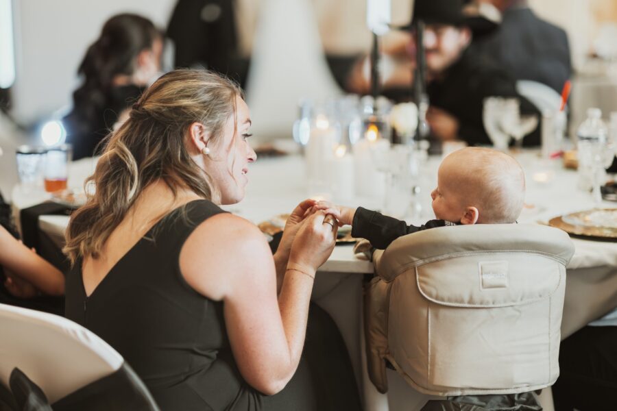 A woman in a black dress interacts with a smiling baby in a high chair during a lively event, surrounded by elegantly set tables and soft candlelight.