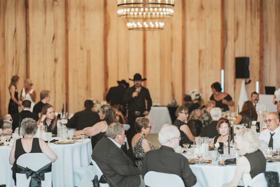 Crowded banquet hall with elegantly dressed guests enjoying a formal dinner, featuring a man in a black cowboy hat speaking at a podium, wooden walls, and a chandelier overhead.