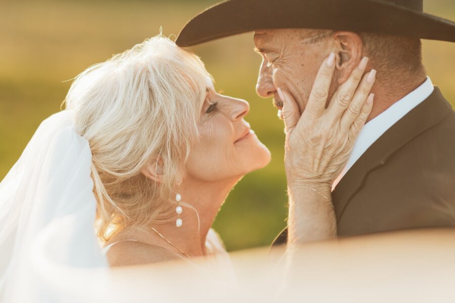 Couple sharing an intimate moment during golden hour, with the bride wearing a veil and pearl earrings, and the groom in a black suit and cowboy hat, set against a soft-focus outdoor background.