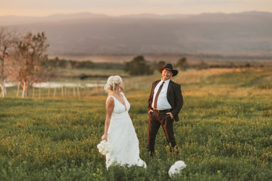 Couple celebrating their wedding in a scenic outdoor setting, with the bride in a white gown holding a bouquet and the groom in a black suit and cowboy hat, surrounded by lush green grass and a picturesque sunset backdrop.